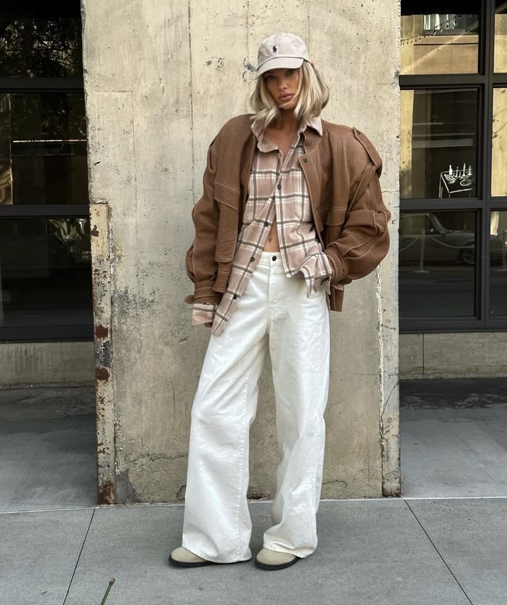 Fashionable woman in brown leather jacket, plaid shirt, and white pants, leaning against a textured wall.