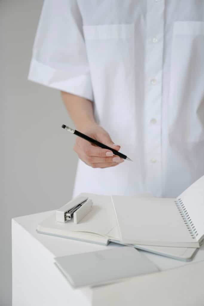 Person in white shirt writing in a notebook with a pencil, near a stapler and another notebook on a white table.