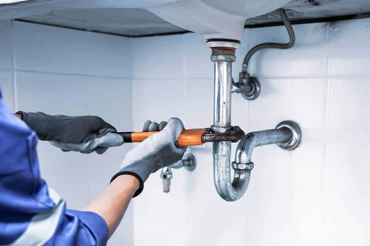 Plumber using a wrench to fix a sink pipe under a kitchen or bathroom basin, wearing protective gloves.