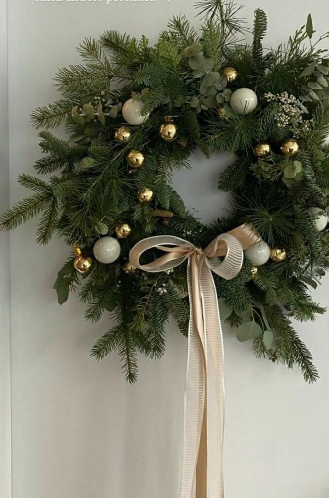 Christmas wreath with pine branches, gold and white ornaments, adorned with a large beige ribbon hanging on a wall.