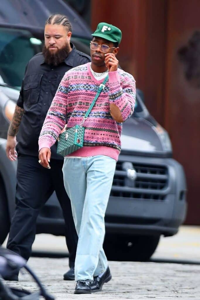 Stylish man in pink patterned sweater and green cap walks in urban setting with bodyguard.