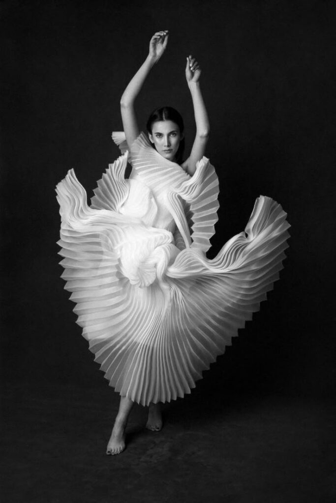 Elegant woman in flowing, pleated dress striking a pose against dark background, showcasing artistic fashion design.