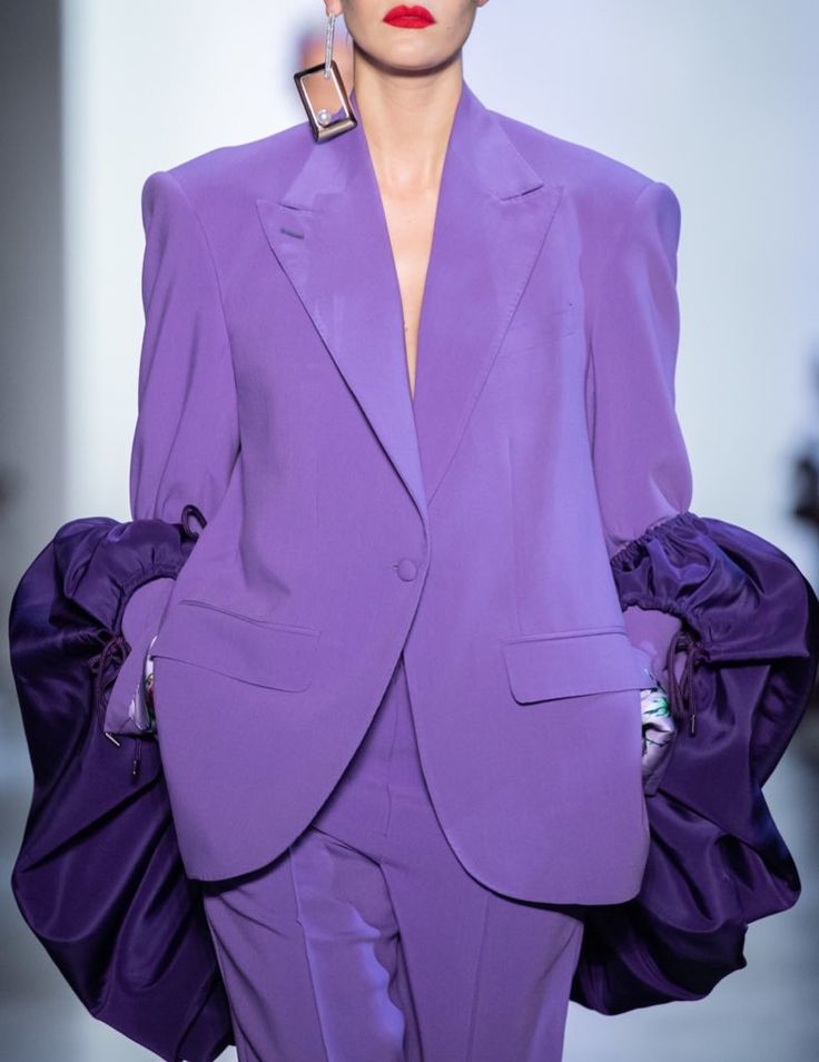 Model in stylish purple suit with bold geometric earrings on runway.