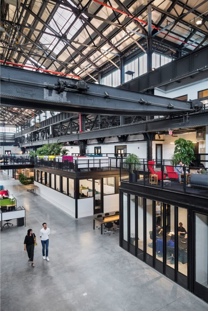 Modern industrial office space with people walking, featuring open plan design and glass-walled meeting rooms.