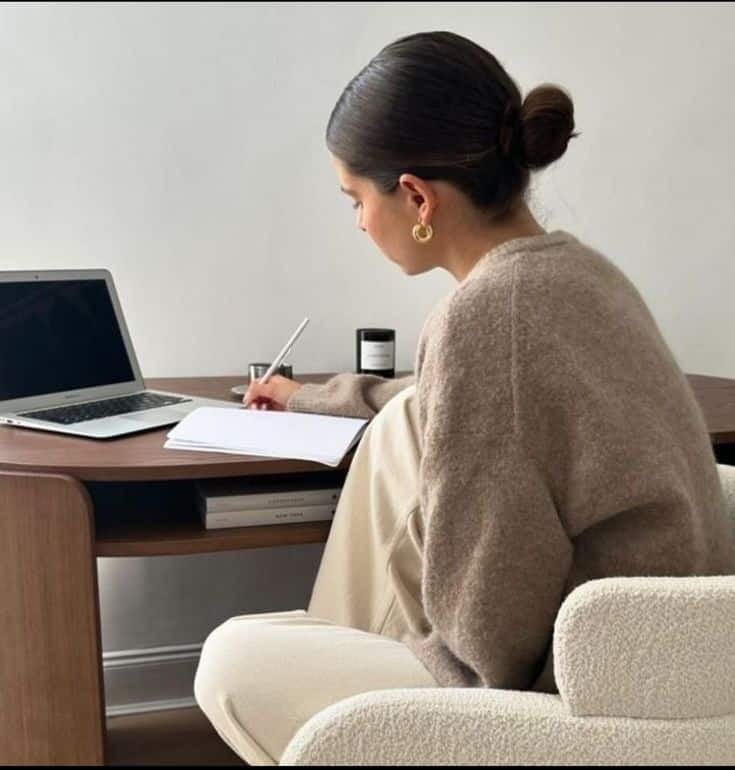 Person writing in a notebook at a desk with a laptop, wearing a cozy sweater.