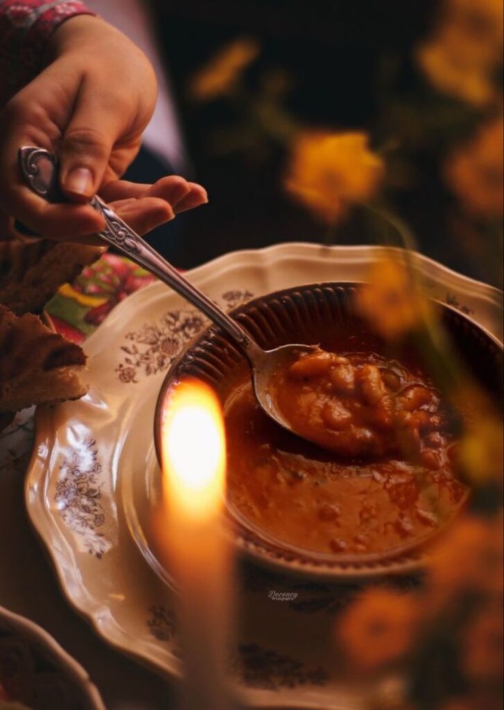 Person serving warm bean soup in a decorative bowl, with candlelight ambiance and vibrant flowers.