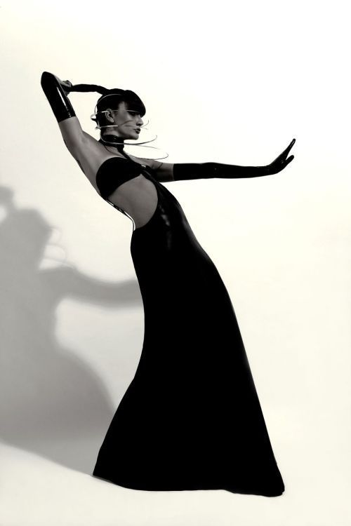 Elegant person wearing a sleek black gown, striking an expressive pose against a plain background.