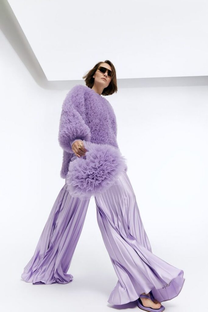 Fashion-forward woman in lavender outfit with fluffy top and flowy pleated pants, wearing sunglasses, on a white backdrop.