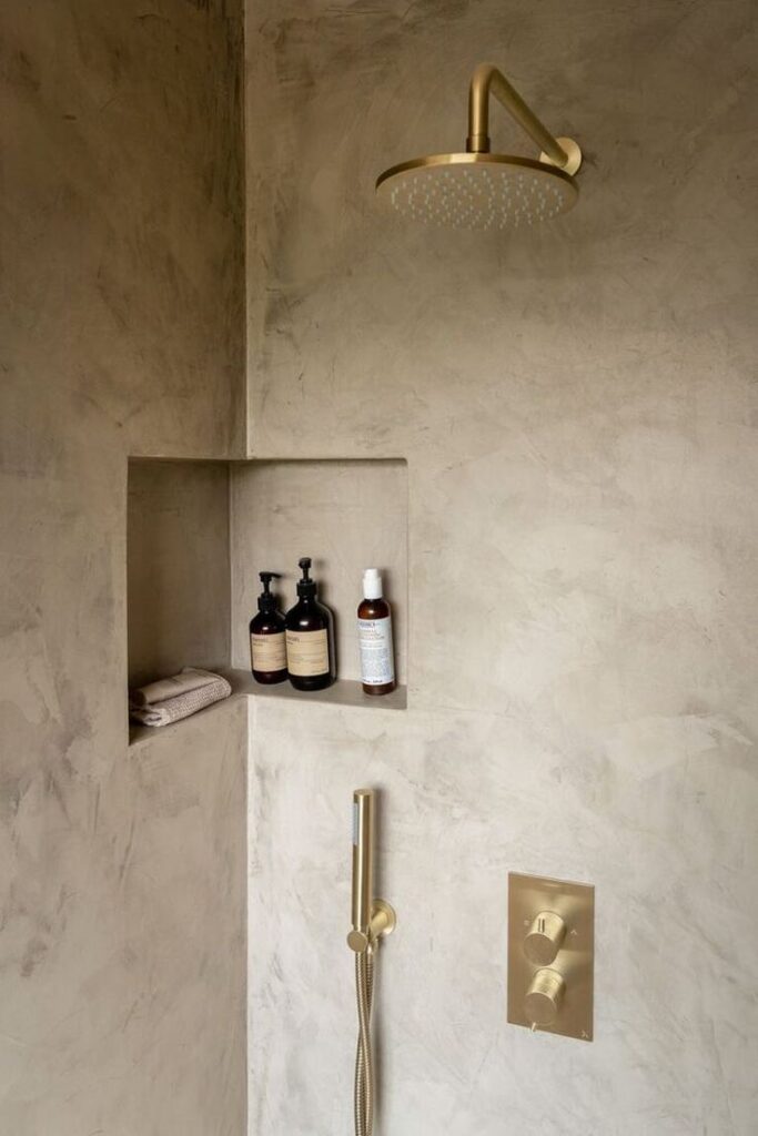 Modern shower with brass fixtures, minimalist niche shelves with toiletries, and neutral stone walls.