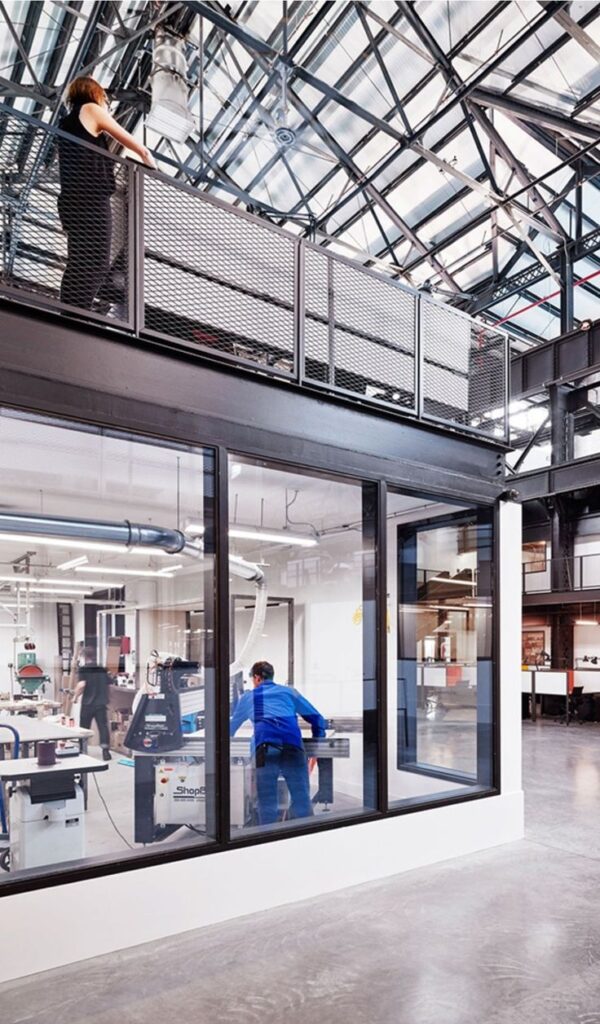 Modern industrial workspace with people working, featuring glass walls, metal beams, and high ceilings.