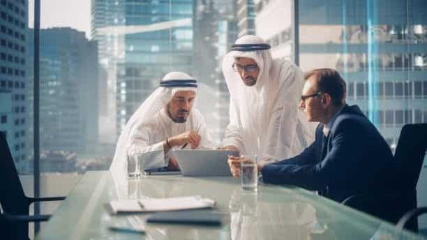 Business meeting with three men in formal attire, collaborating on a laptop in a modern glass office with a cityscape view.