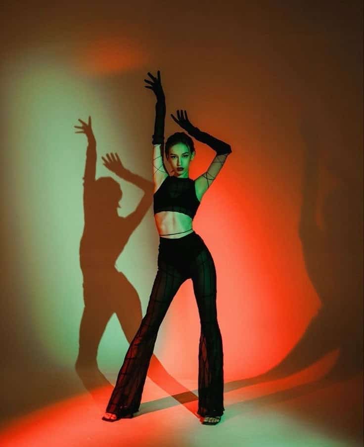 Dancer poses dramatically in black outfit against colorful red and green lighting backdrop, casting dynamic shadows.