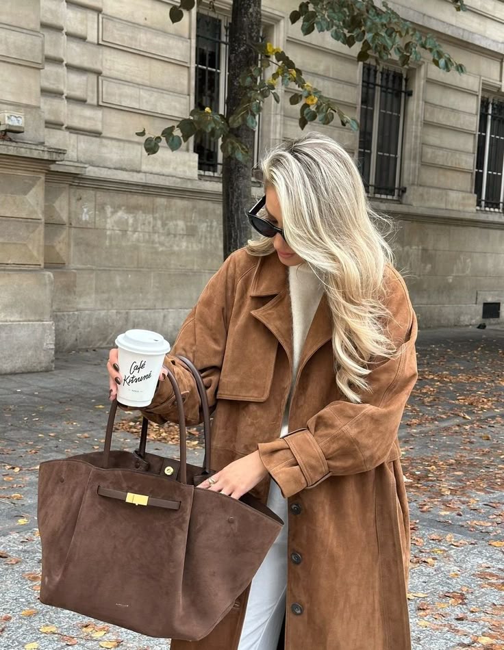 Woman in stylish brown coat holding coffee and handbag on a city street.