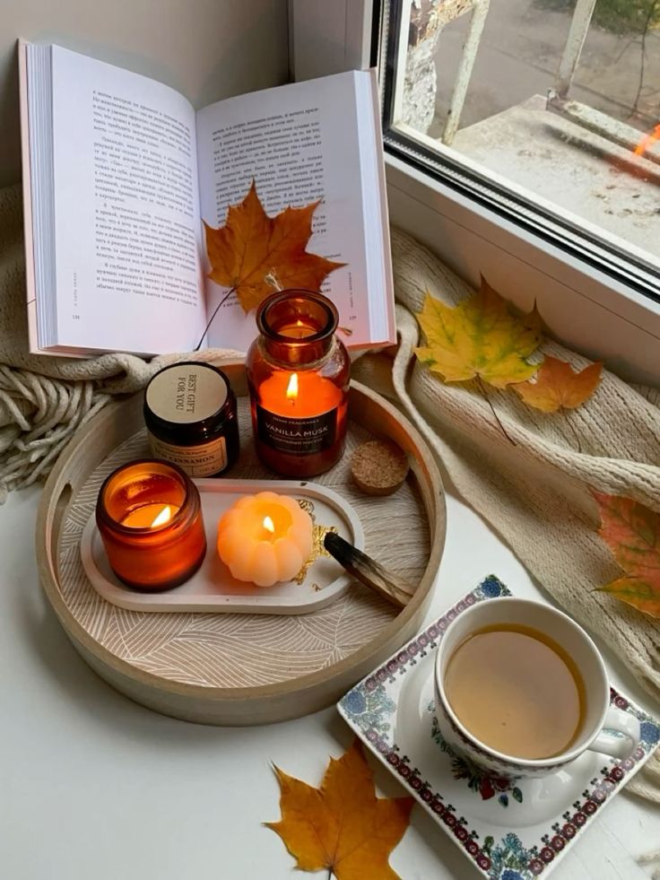 Cozy autumn scene: Open book, candles, fall leaves, and tea by the window on a wooden tray.