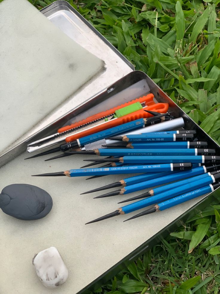 Box of sketching pencils, erasers, and a cutting tool on grass, ideal for artists drawing outdoors.