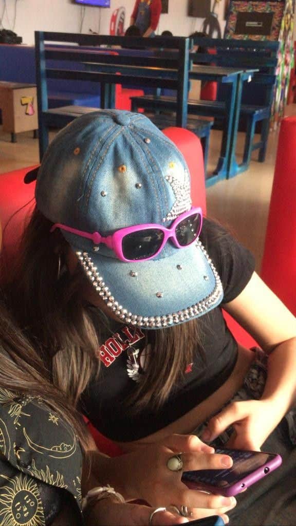 Young person wearing a denim cap and sunglasses, focused on a smartphone in a casual setting.
