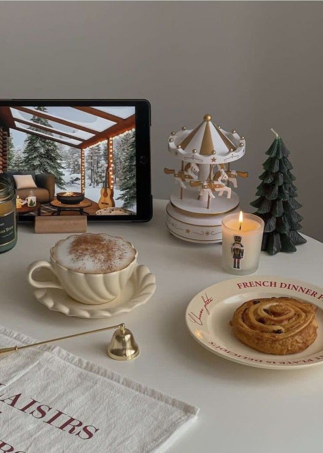 Cozy setup with cappuccino, pastry, candle, carousel, tablet showing winter scene, and holiday decor on a white table.