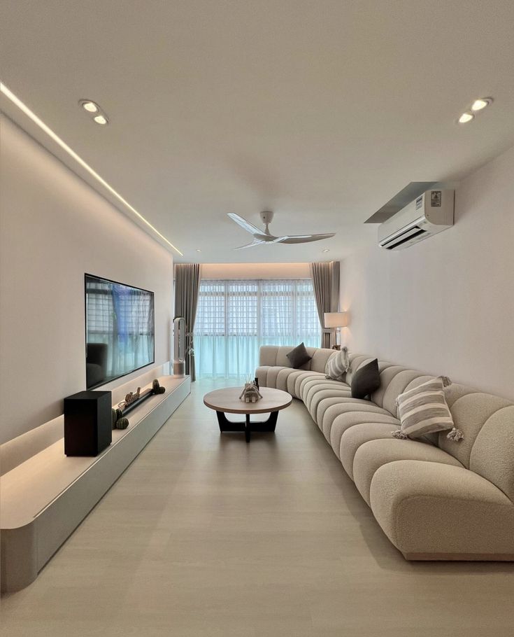 Modern living room with beige sectional sofa, wall-mounted TV, and natural light from large windows with sheer curtains.