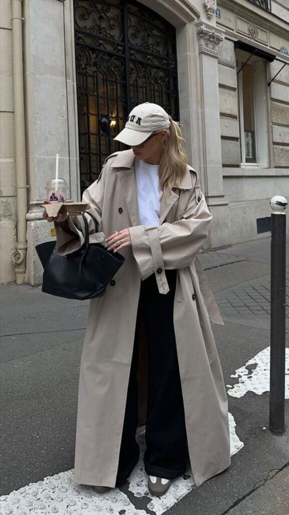 Stylish woman in trench coat and cap carrying coffee and bag on city street.