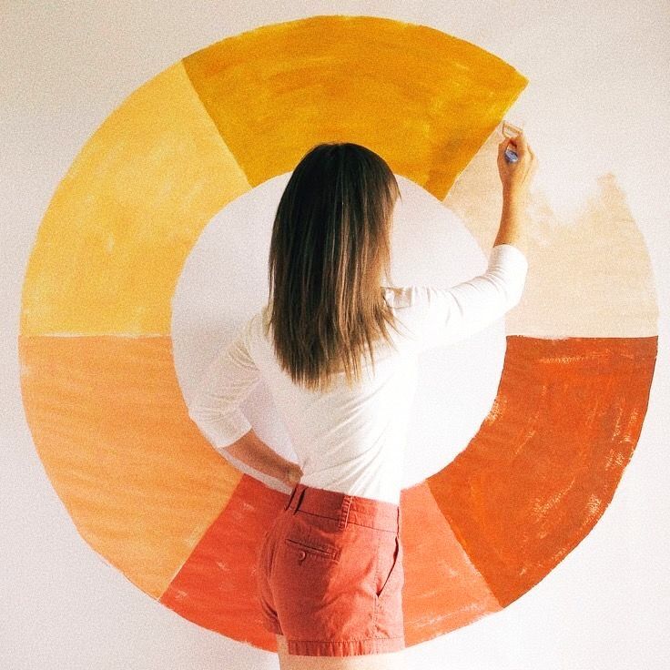 Woman painting colorful circle mural on white wall, wearing red shorts and white shirt. Creative home art project.