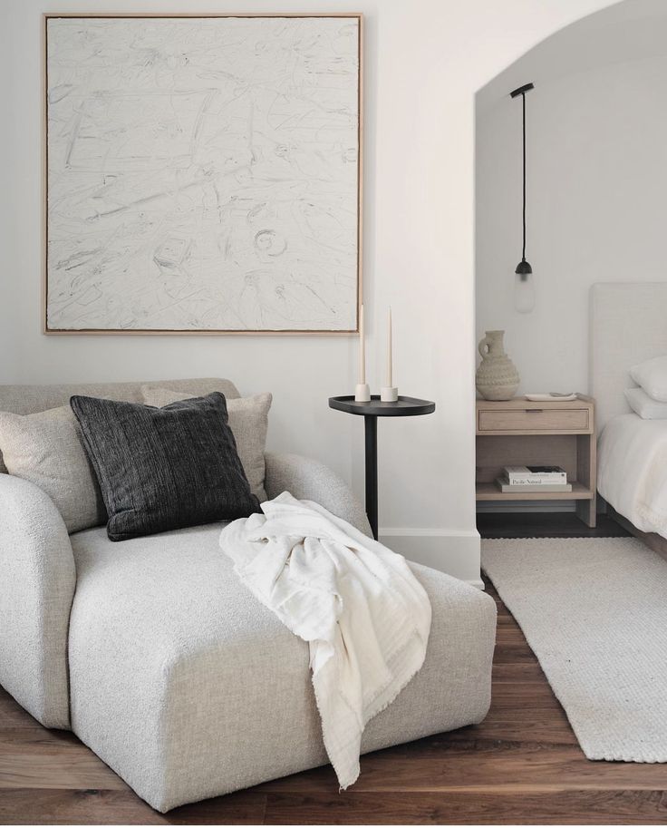 Minimalist living space with modern gray chair, black pillow, and abstract wall art, next to a bedroom nook.