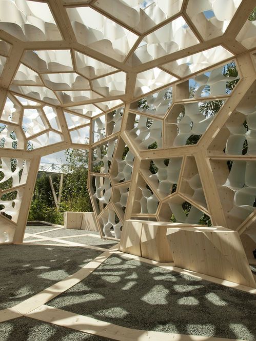 Modern architectural pavilion with geometric wooden structure, dappled sunlight, and a view of trees.