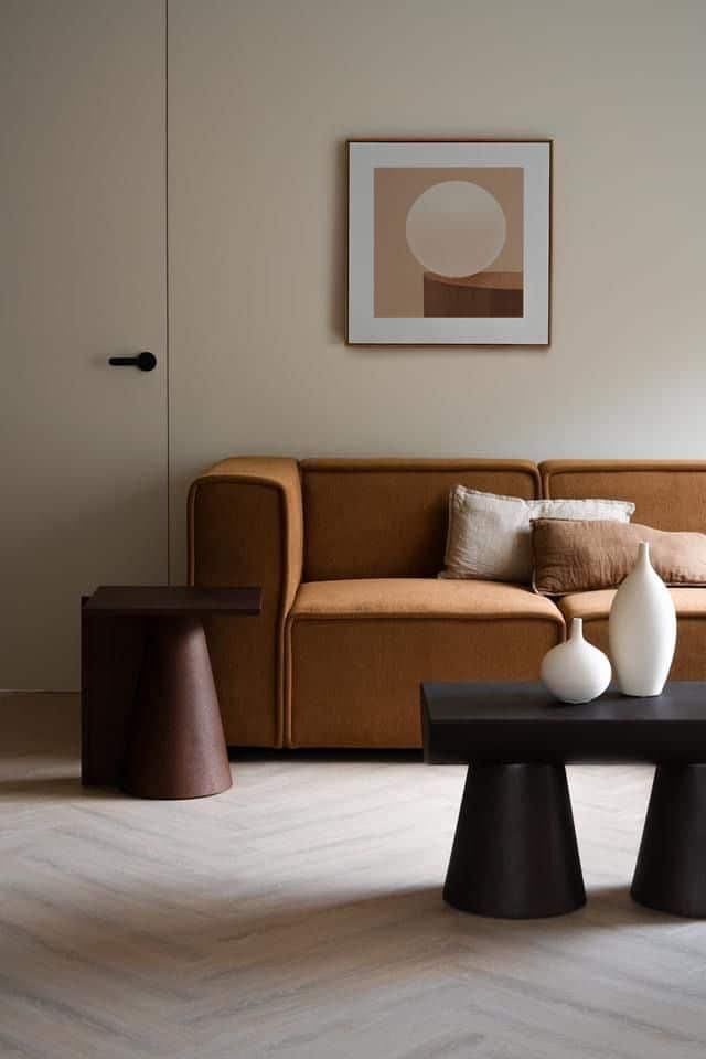 Minimalist living room with brown sofa, modern tables, and abstract wall art in neutral tones.