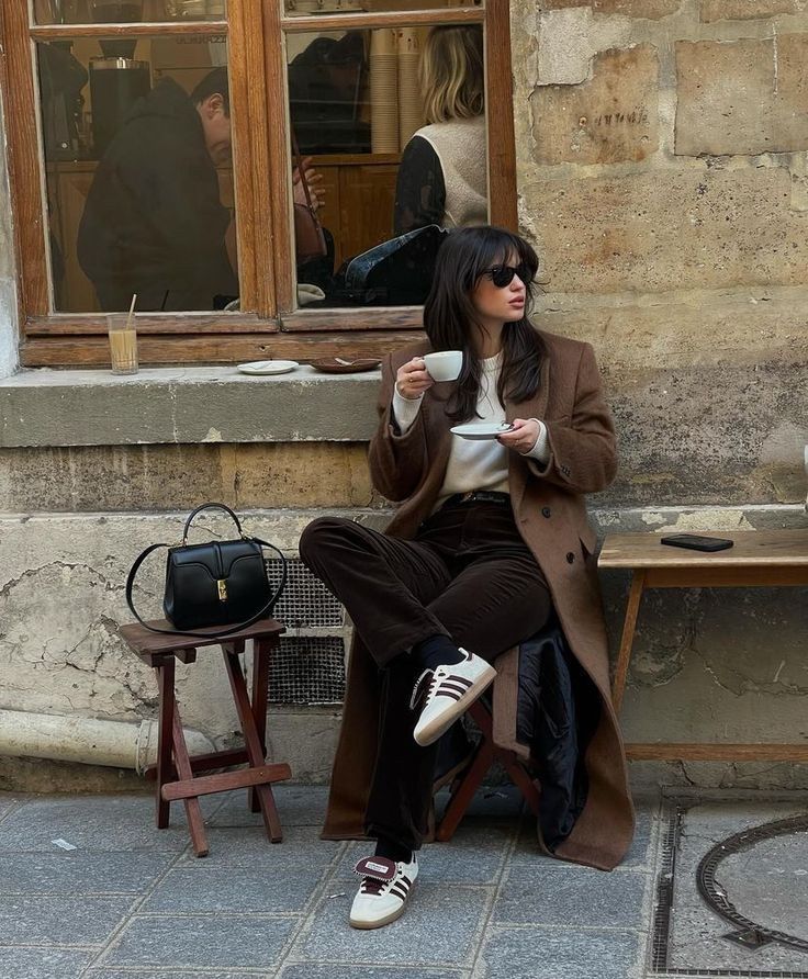 Stylish woman in sunglasses enjoys coffee at an outdoor café, wearing a brown coat and sneakers.