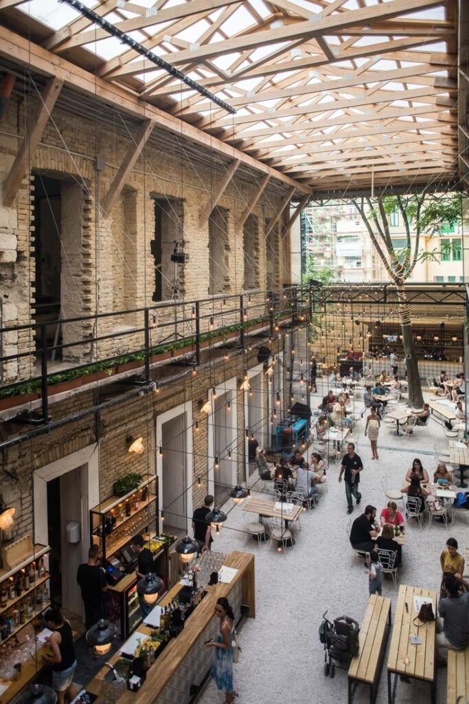 Airy café with people dining under wood-beam ceiling, featuring rustic decor and abundant natural light.