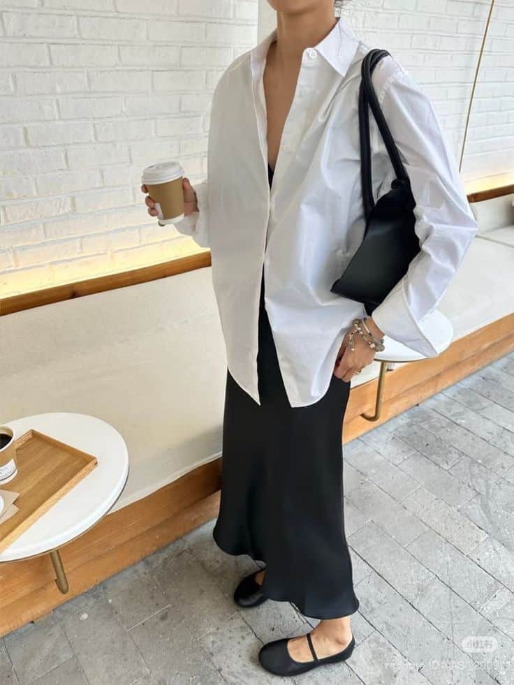 Stylish woman in a white shirt, black skirt, and flats holding a coffee cup, accessorized with a black handbag.