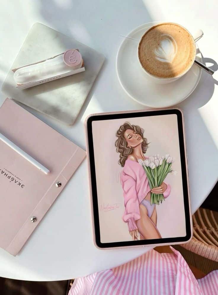 Digital art on tablet with cappuccino, pink notebook, and pen on a white table. Cozy, creative workspace.