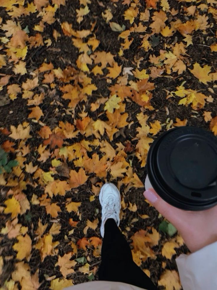 Walking through autumn leaves while holding a coffee cup, person in white sneakers and black pants.
