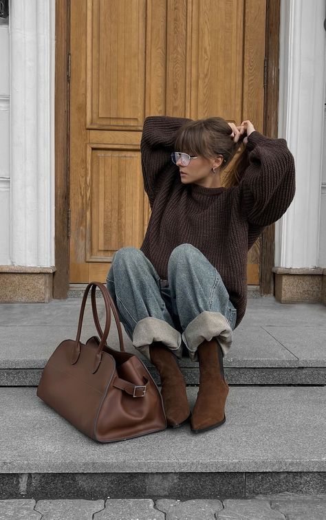 Woman in cozy sweater and jeans sitting on steps with brown boots and matching bag, styling hair.