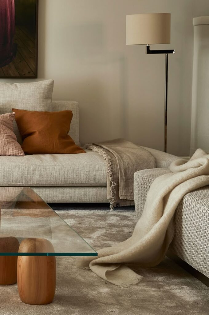 Modern living room with beige sofa, cozy cushions, a floor lamp, and a glass coffee table on a plush carpet.