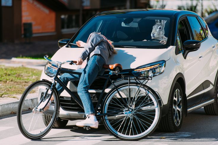A person on a bicycle collides with a white car driven by someone wearing a rabbit mask in a humorous scene.