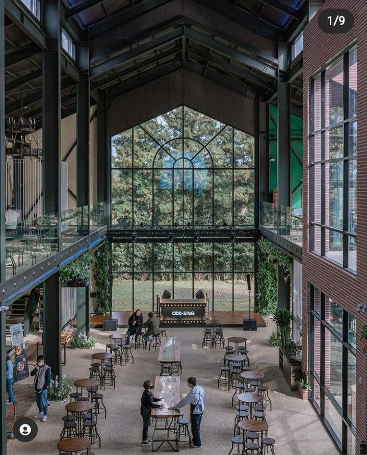 Modern industrial interior with high ceilings, large windows, tables, and people socializing in a spacious lounge area.