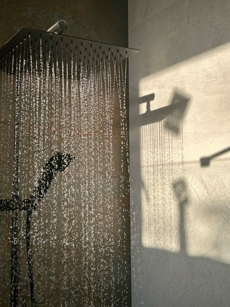 Modern rainfall showerhead with flowing water in a sunlit bathroom, casting shadows on the wall.