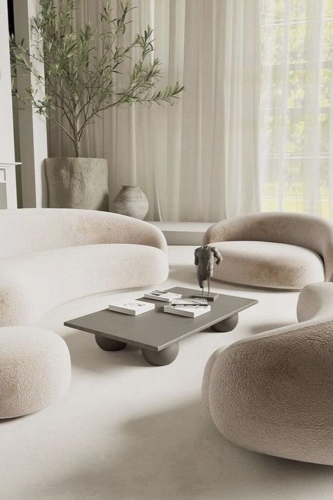 Modern minimalist living room with neutral curved sofas, low table, potted plant, and elegant decor.