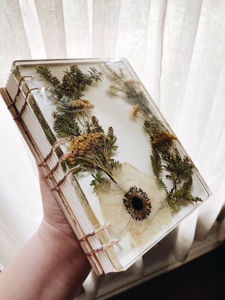 Hand holding a handmade journal with pressed flowers under resin, against a soft, light-filtering curtain background. handmade items