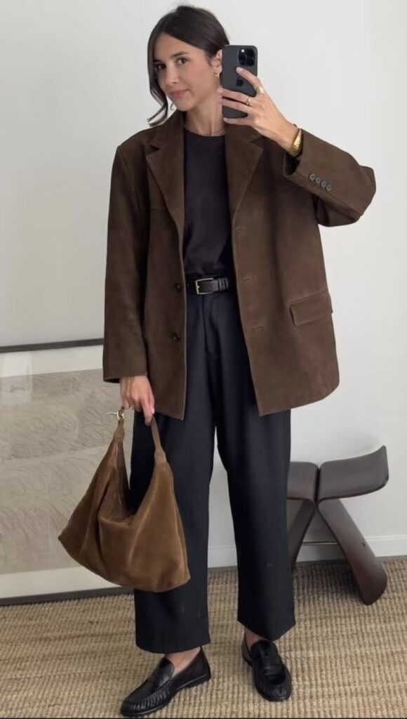 Fashionable woman in brown blazer and black pants taking a mirror selfie with a brown handbag.
