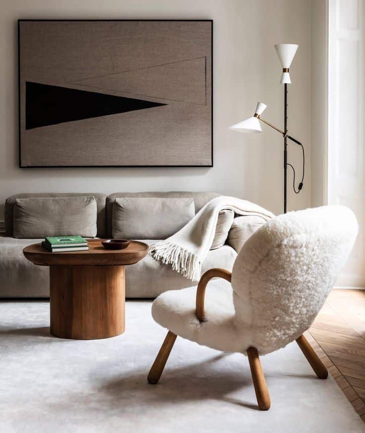 Modern living room with cozy armchair, wooden table, abstract wall art, and floor lamp. Neutral tones create warmth.