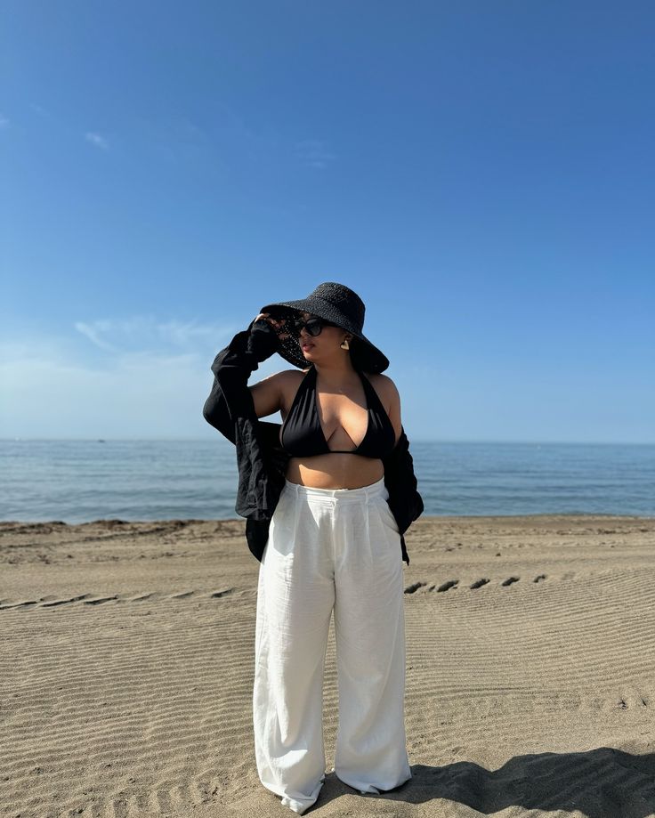 Woman in sun hat and stylish beachwear stands on sandy beach under clear blue sky.
