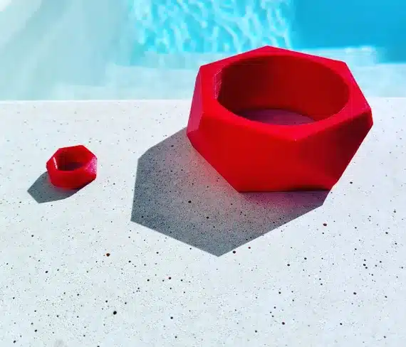 Red geometric bracelet and ring on a white surface near a pool, casting distinct shadows in the sunlight.