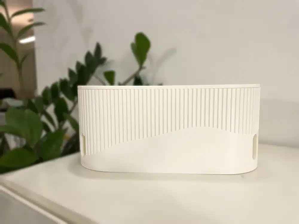 White textured cable management box on a desk with green plants in the background.