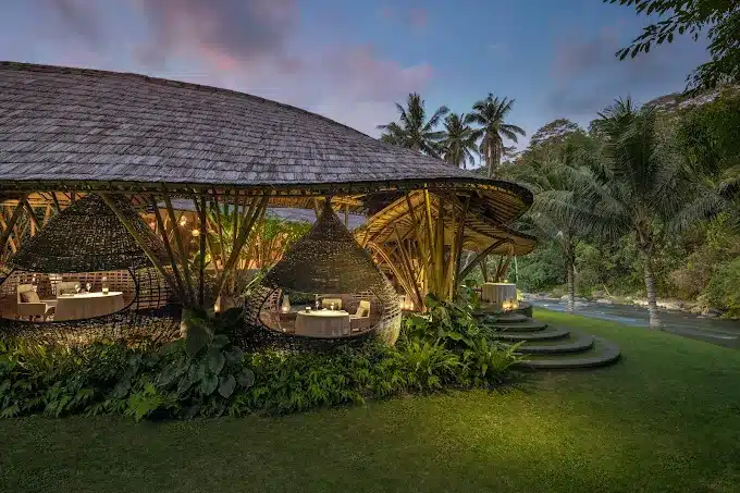 Rustic bamboo restaurant by riverside with lush greenery and ambient lighting, set against a tropical evening backdrop.