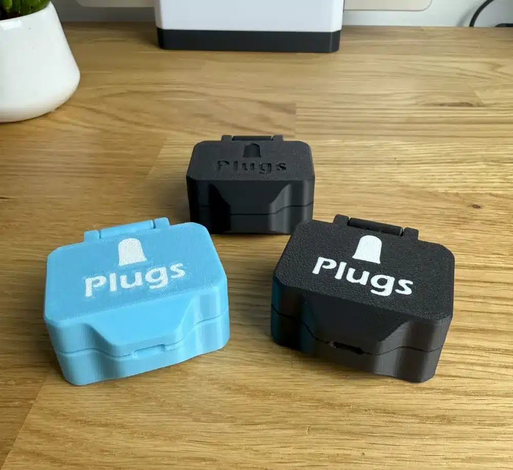 Three portable earplug cases on a wooden surface, one in blue and two in black, labeled Plugs with a bell icon.