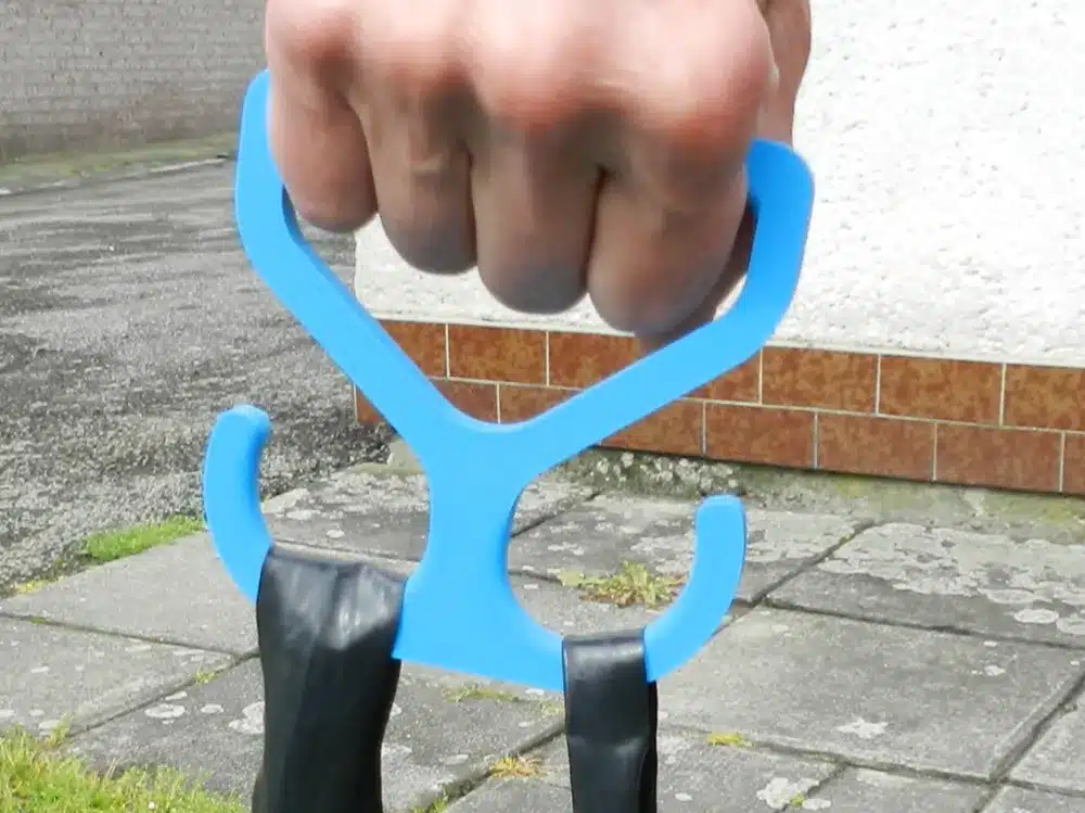 Hand holding blue hook tool gripping black rubber bands on concrete pavement.