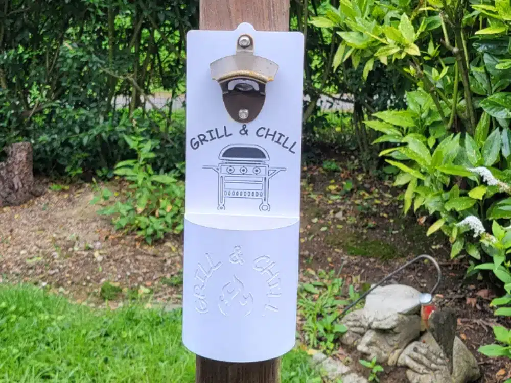 Wall-mounted bottle opener with Grill & Chill design in a garden setting. Perfect for outdoor BBQs and parties.