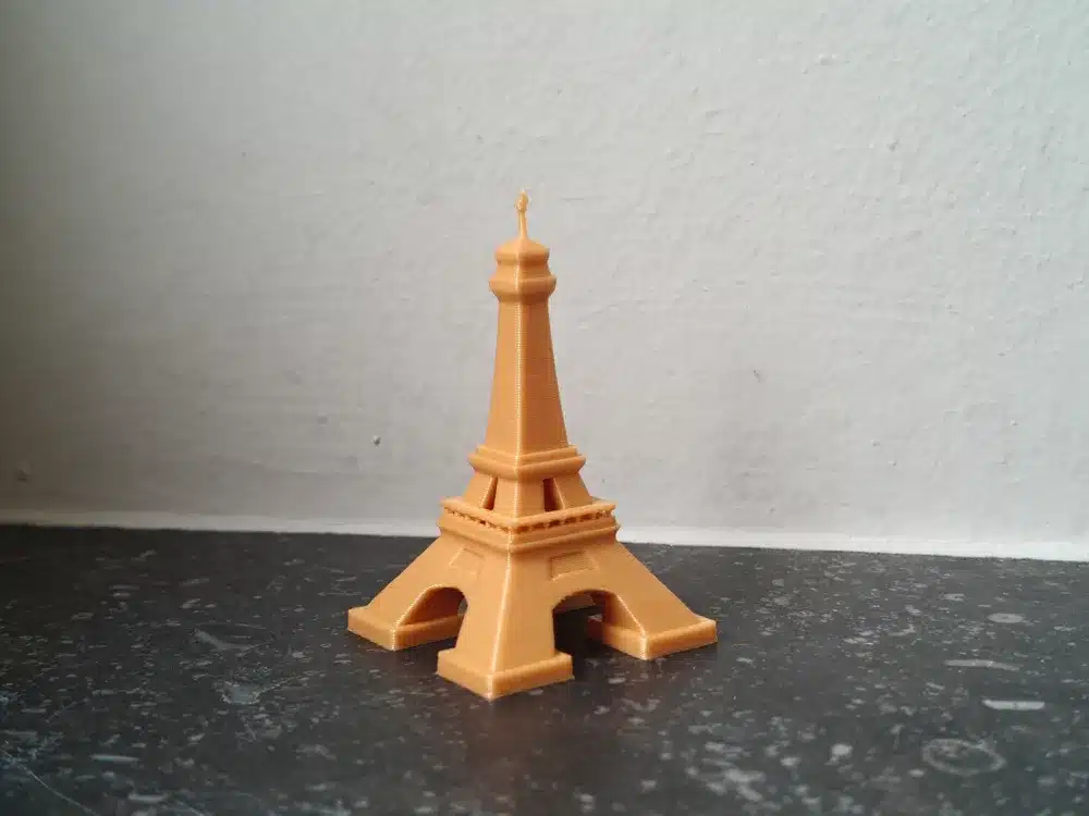 3D-printed Eiffel Tower model in orange on a dark surface, against a light wall background.