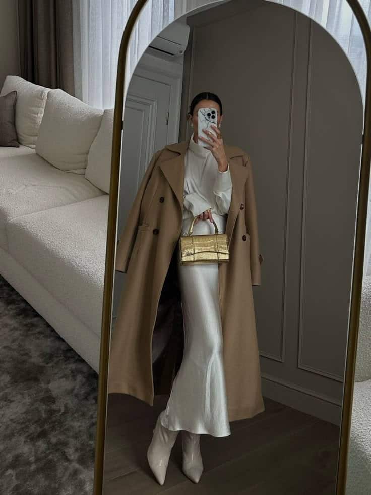 Stylish woman in long camel coat and white dress taking mirror selfie with gold handbag and smartphone.